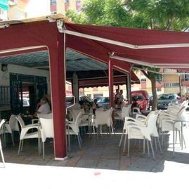 Terraza de cafetería con asientos bajo toldos rojos, sillas y mesas blancas.