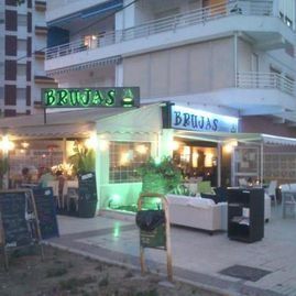 Cafetería al aire libre llamada “Brujas” con letrero iluminado en verde y mesas afuera al atardecer.