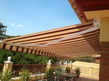 Patio con toldo retráctil a rayas sobre muebles de exterior y plantas en macetas bajo un cielo azul.