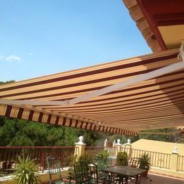 Patio sombreado con toldo retráctil a rayas, mesas, sillas y macetas bajo un cielo azul despejado.
