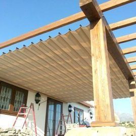 Pérgola de madera con techo de listones que da sombra sobre un patio, junto a una casa blanca con escaleras cerca.