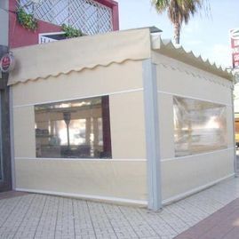 Pequeño quiosco de café al aire libre de color beige con toldo y grandes ventanales en una acera.
