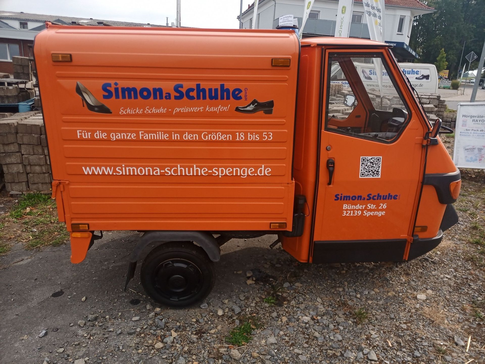 Orangefarbener Lieferwagen von Simon Schuhe, geparkt auf Schotter, Seitenansicht mit Firmenlogo und Website.