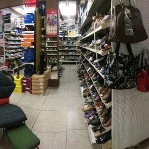 Schmaler Schuhladengang mit Regalen voller Schuhe und Handtaschen, gefliestem Boden und gestapelten Kartons.