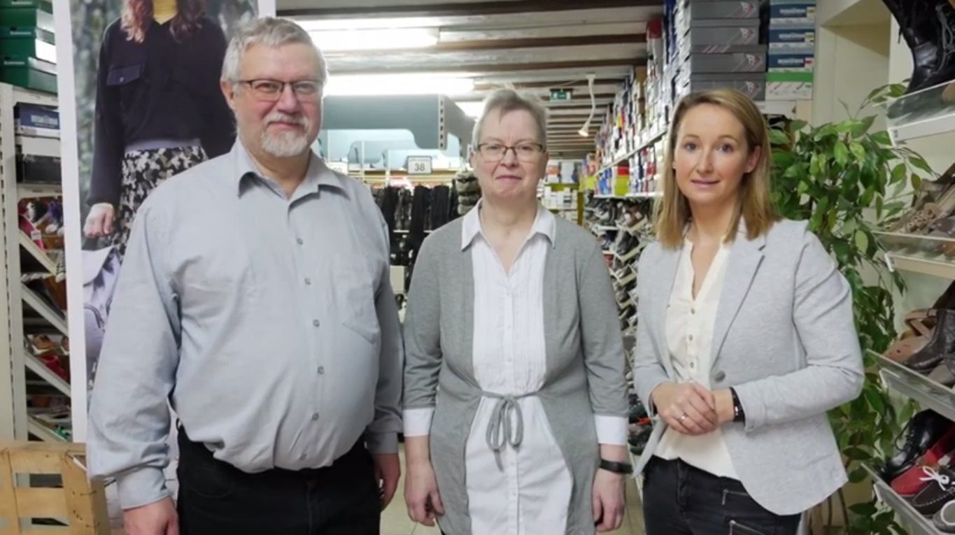 Drei Personen stehen in einem Ladengang, blicken in die Kamera, hinter ihnen befinden sich Regale mit Waren.