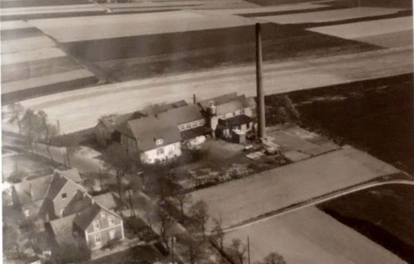 Luftaufnahme eines ländlichen Bauernhauses mit Nebengebäuden neben Feldern, mit einem hohen Schornstein in der Nähe der Straße