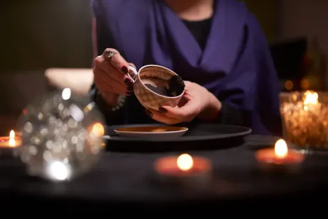 Persona vertiendo posos de café de una taza en un platillo, rodeada de velas y una bola de cristal.