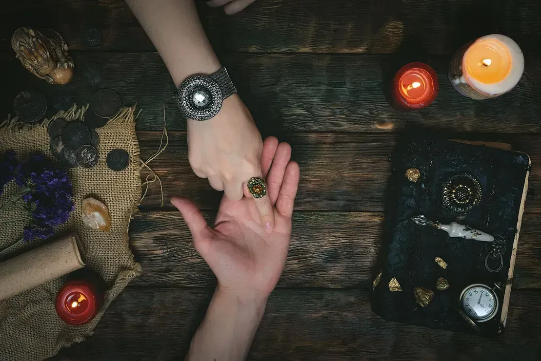 Manos unidas durante la lectura de la palma; velas, libro y accesorios sobre una mesa de madera.