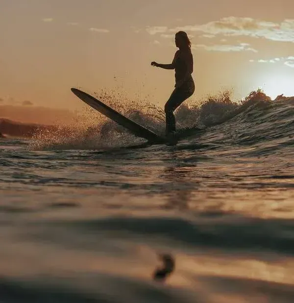 Silhouette d’un surfeur glissant sur une vague au coucher du soleil, avec la lumière dorée qui se reflète sur l’eau.