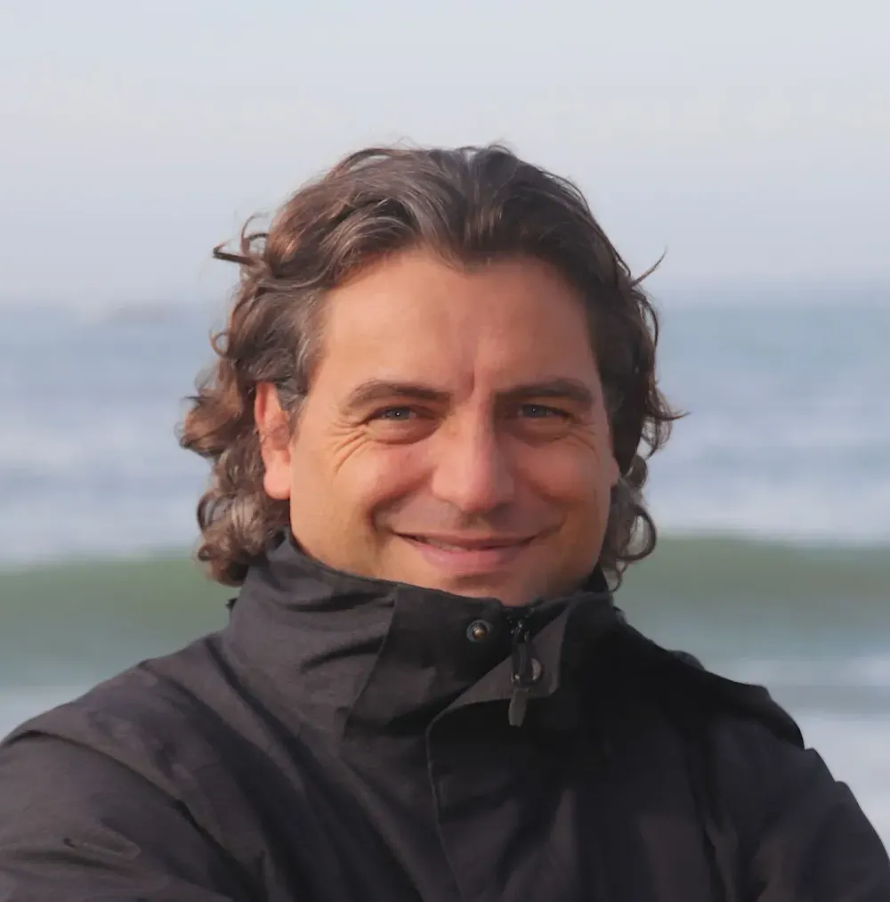 Personne souriante portant une veste sombre en extérieur, avec des vagues en arrière-plan