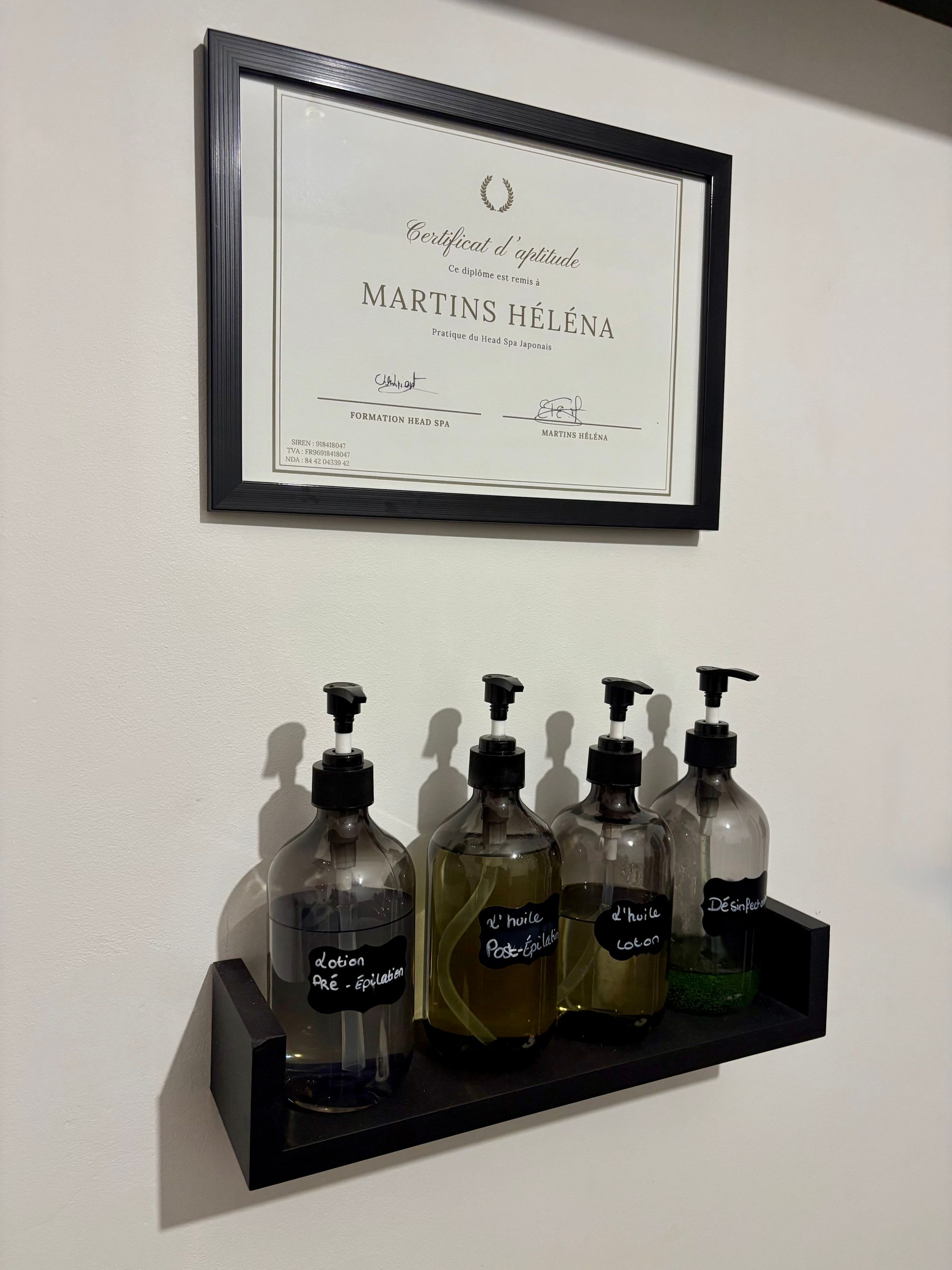 Un certificat encadré est accroché au-dessus d'une petite étagère noire où reposent quatre flacons-pompes en verre.