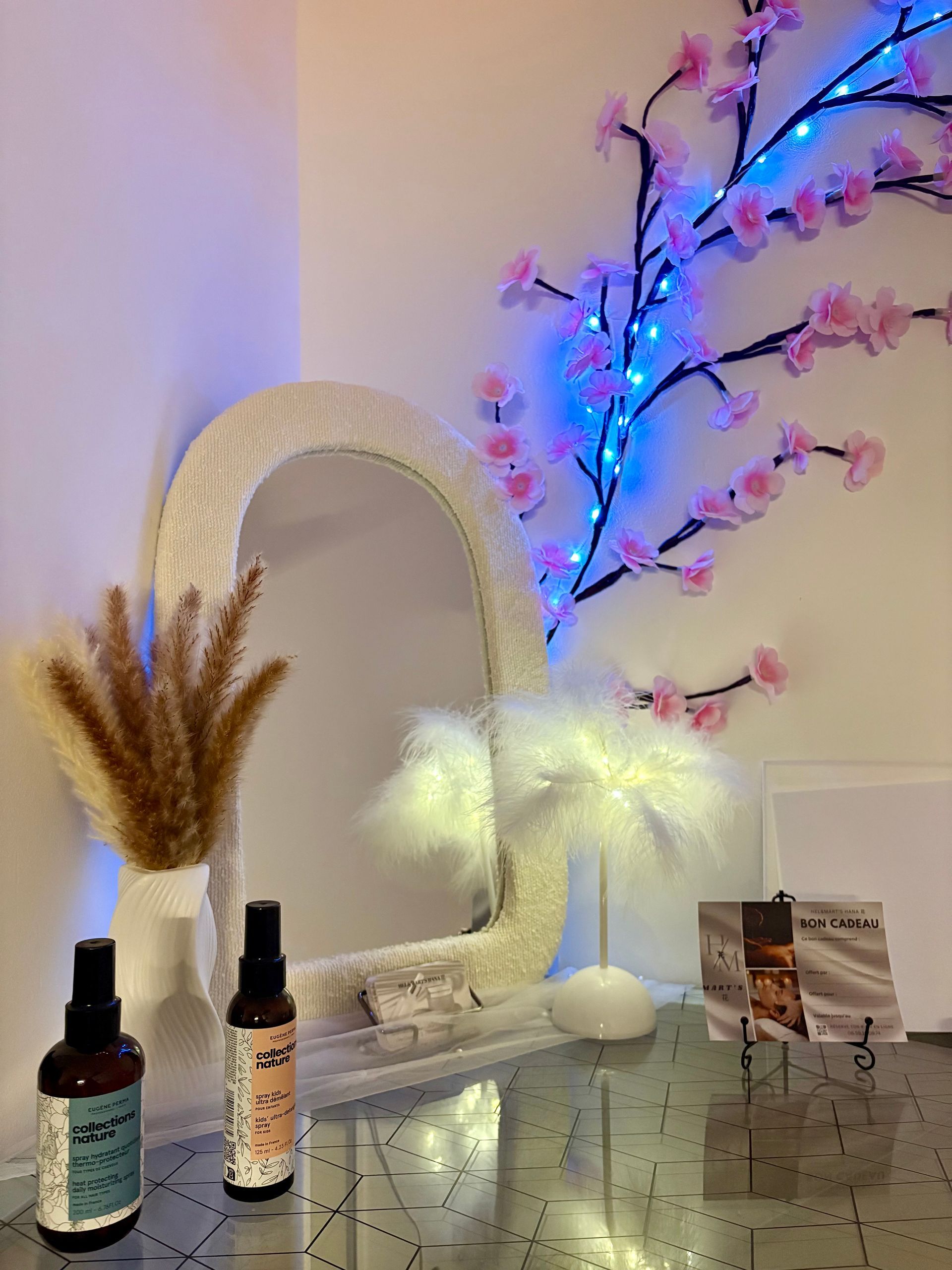 Un bureau avec un miroir, deux flacons vaporisateurs, une lampe à plumes lumineuse et une branche fleurie.