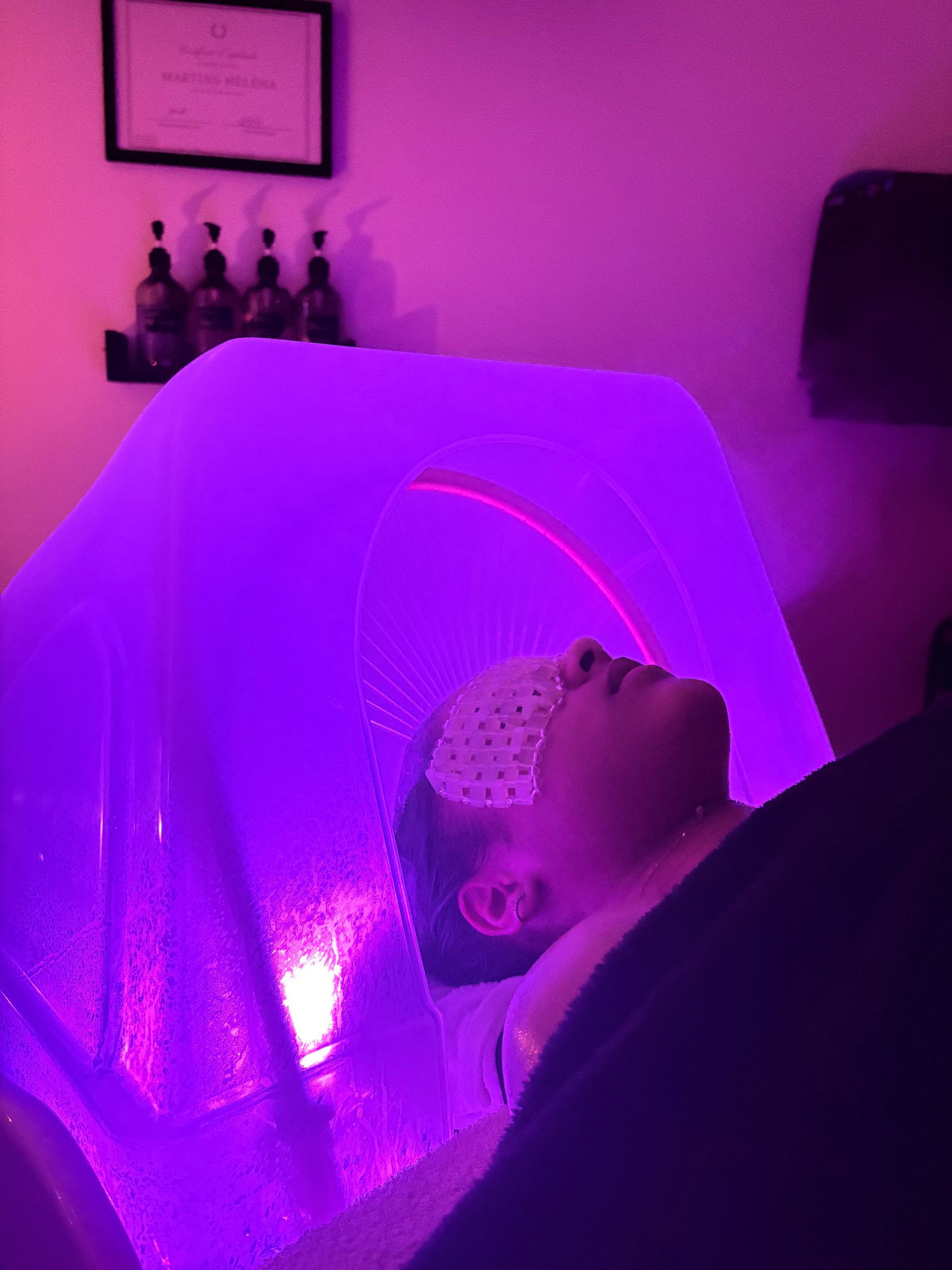 Une personne allongée sur un lit de spa sous un dôme de luminothérapie violette, avec des protections oculaires.