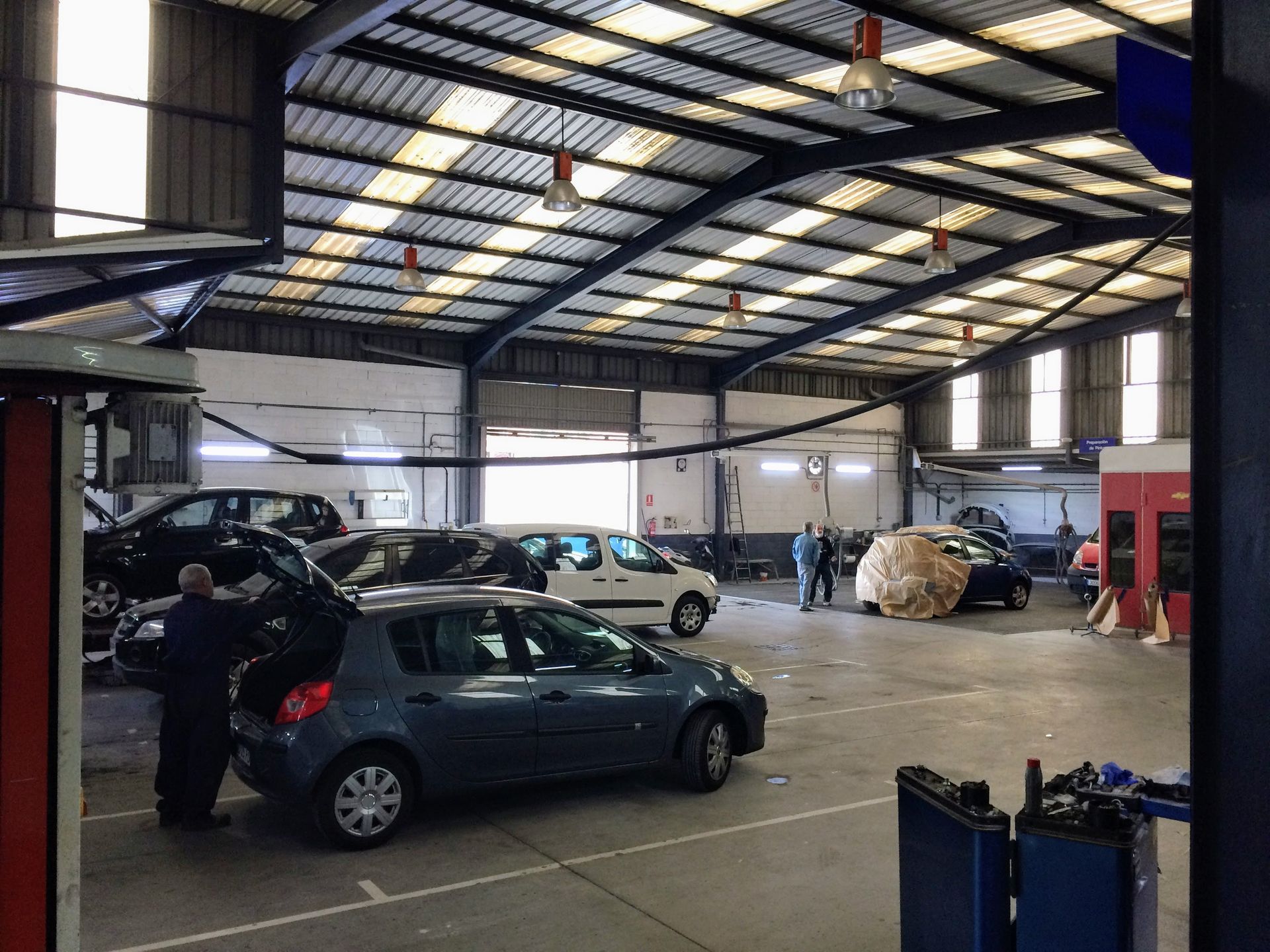 Un coche negro en un taller de reparación con un coche azul al lado, sobre un suelo de rejilla metálica.