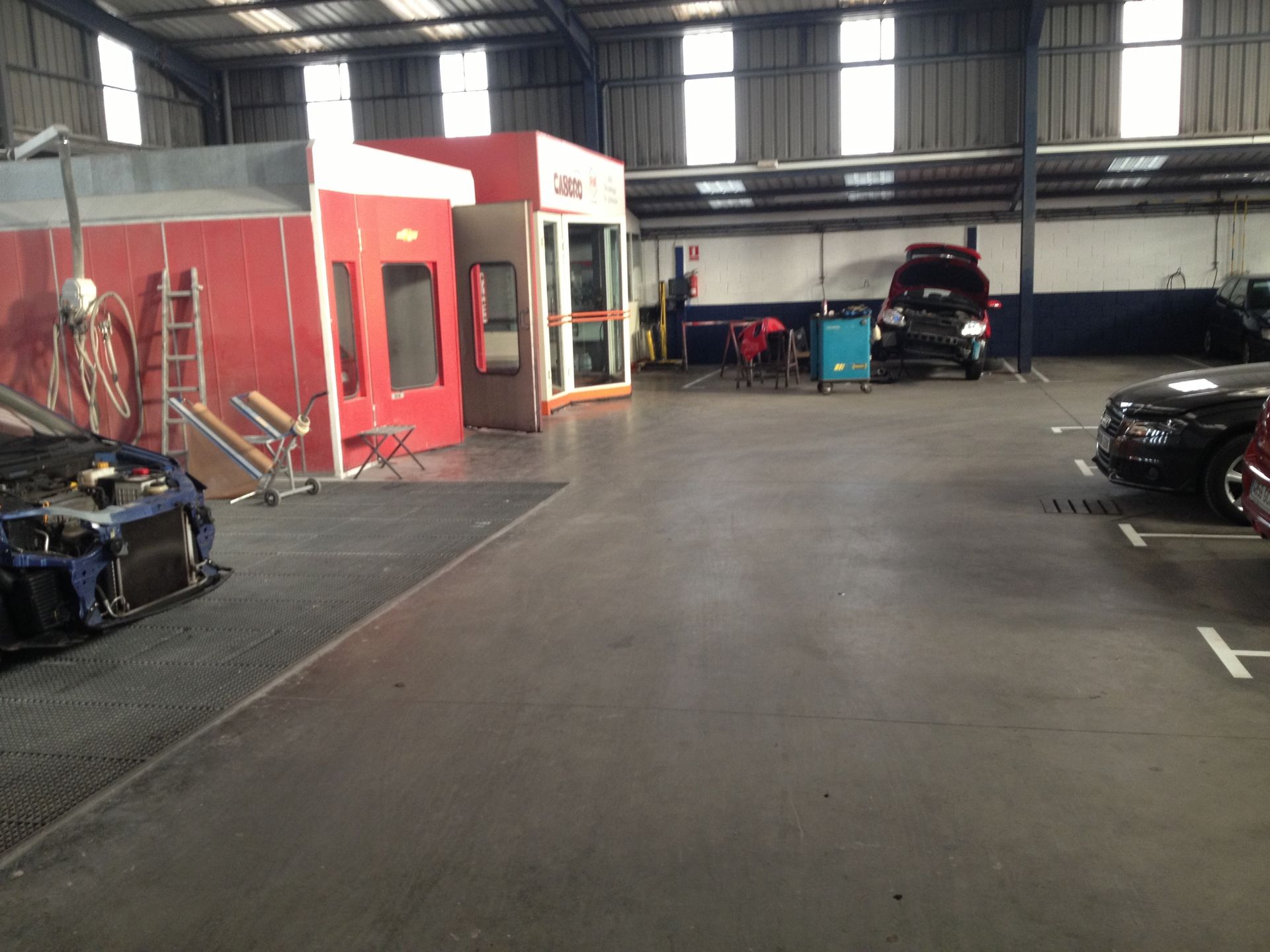 Dentro de un taller de reparación de automóviles, hay una cabina de pintura roja cerca de un automóvil al que le han quitado la parte delantera.