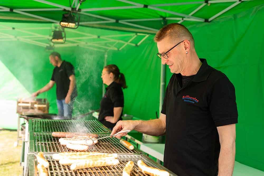 Ein Mann grillt unter einem grünen Zelt Würstchen.