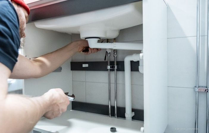 Eine Person arbeitet unter einem Waschbecken und justiert die Wasserleitungen in einem Badezimmerschrank.