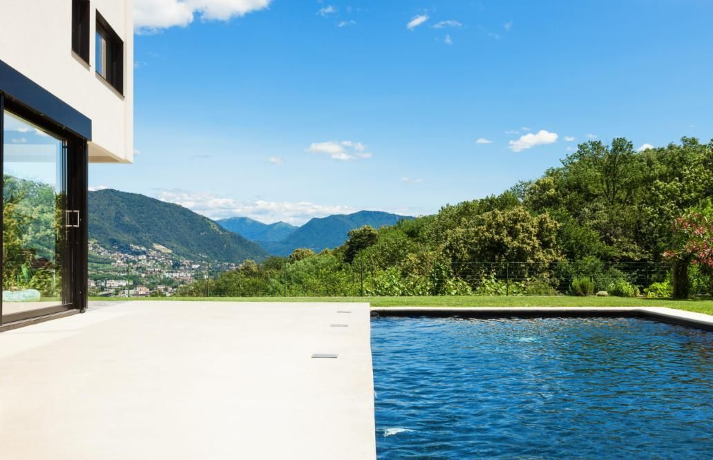 Una casa moderna con piscina domina una ladera verde y una cordillera azul bajo un cielo brillante y soleado.