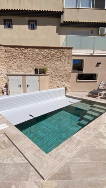 Una piscina rectangular con una cubierta blanca parcialmente cerrada en un patio de piedra junto a una casa