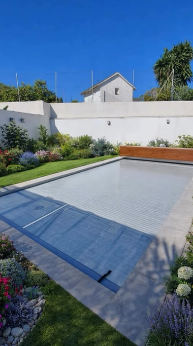 Una piscina rectangular con cubierta de listones en un patio trasero, rodeada de pavimento de piedra