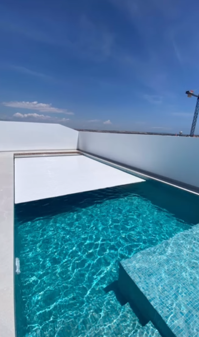 Una piscina rectangular con agua azul y una cubierta blanca parcial, con un cielo azul brillante de fondo.