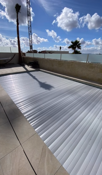 Una piscina cubierta con una lona de seguridad automática de listones blancos en un patio soleado bajo un cielo azul.