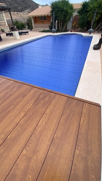 Una piscina rectangular cubierta con una lona de seguridad azul de listones, bordeada por una terraza de piedra clara