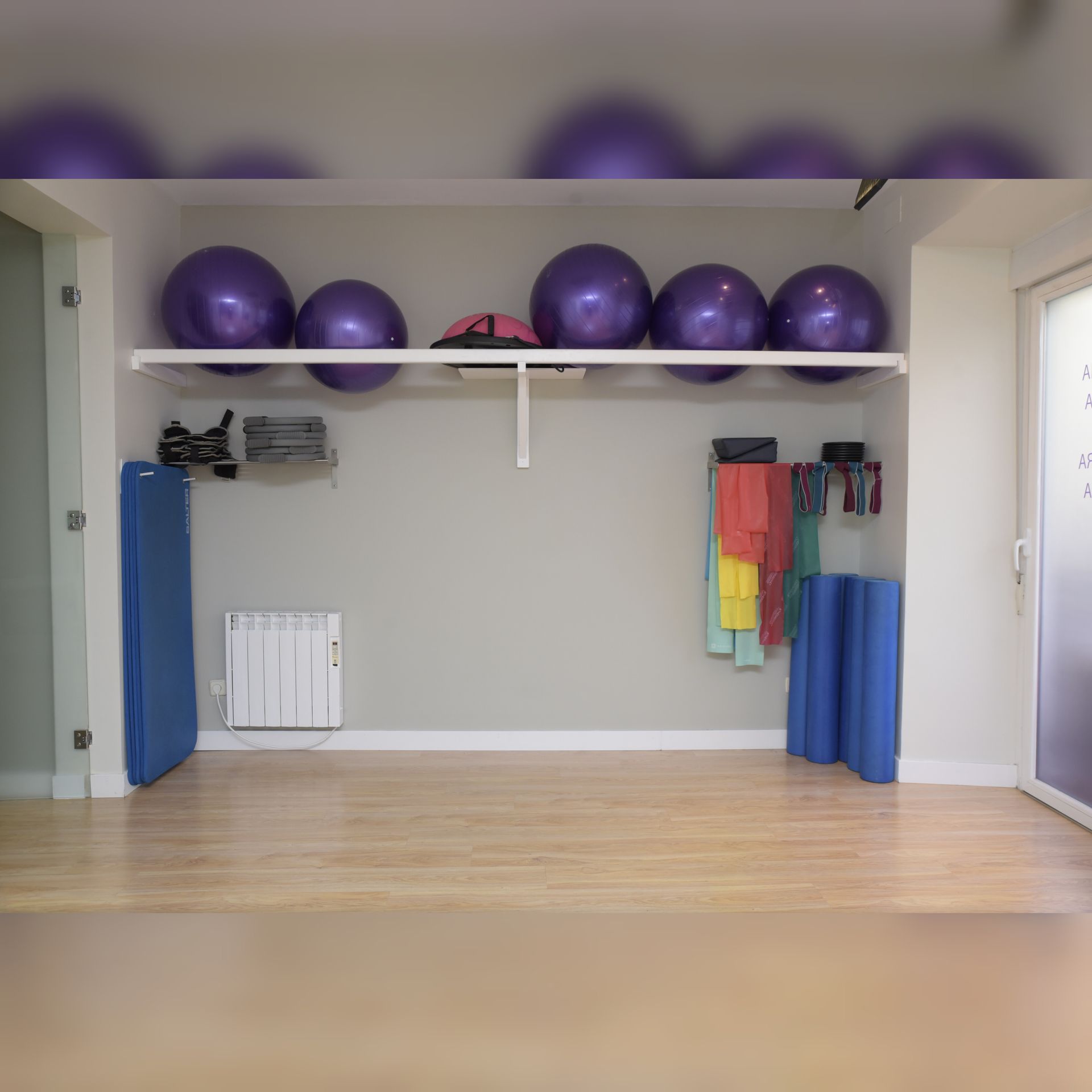 Interior de gimnasio con pelotas de ejercicio de color morado en un estante, colchonetas y otros equipos en la pared, suelo