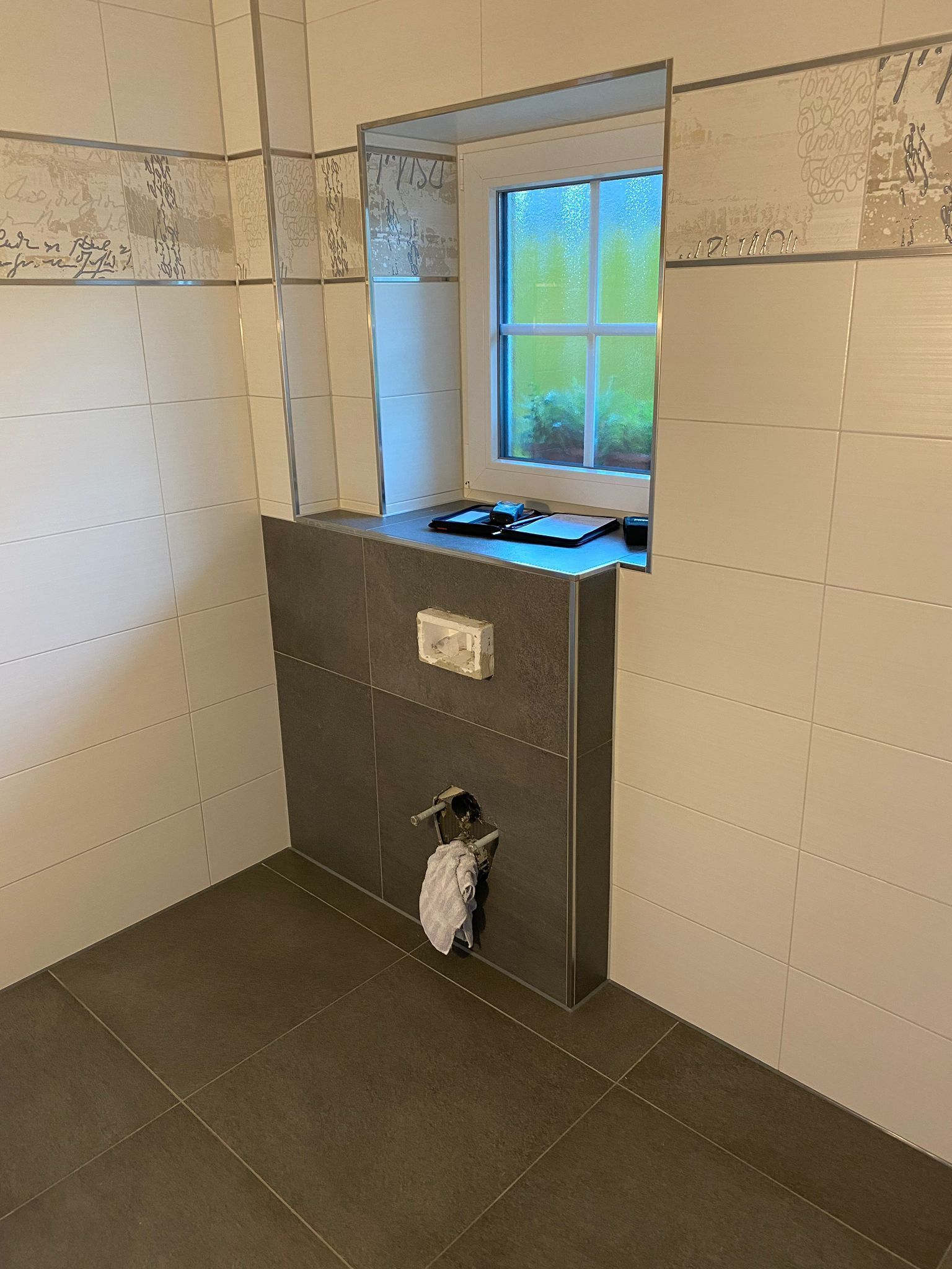 Badezimmer mit einem Fenster über einer grauen Toilettenwand. Wände und Boden sind weiß und grau gefliest.