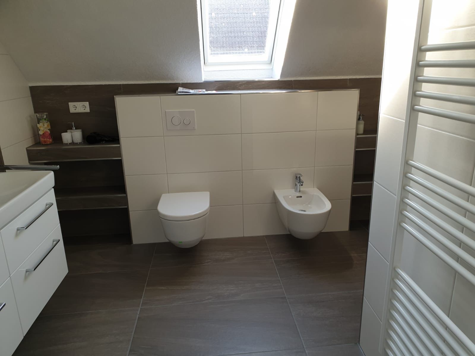 Badezimmer mit weißer Toilette und Bidet, braunem Fliesenboden und einem Dachfenster.
