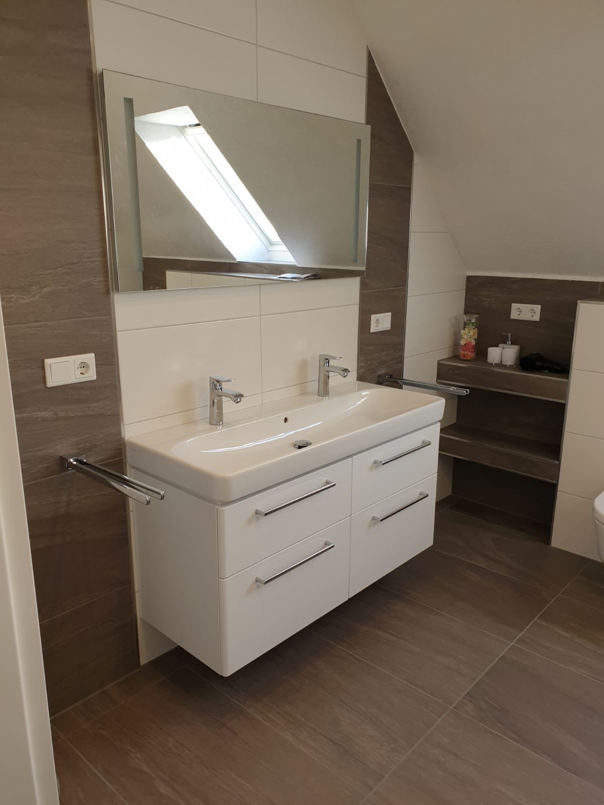 Badezimmer mit weißem Waschbecken, Chromarmaturen, großem Spiegel und braun gefliesten Wänden und Boden.