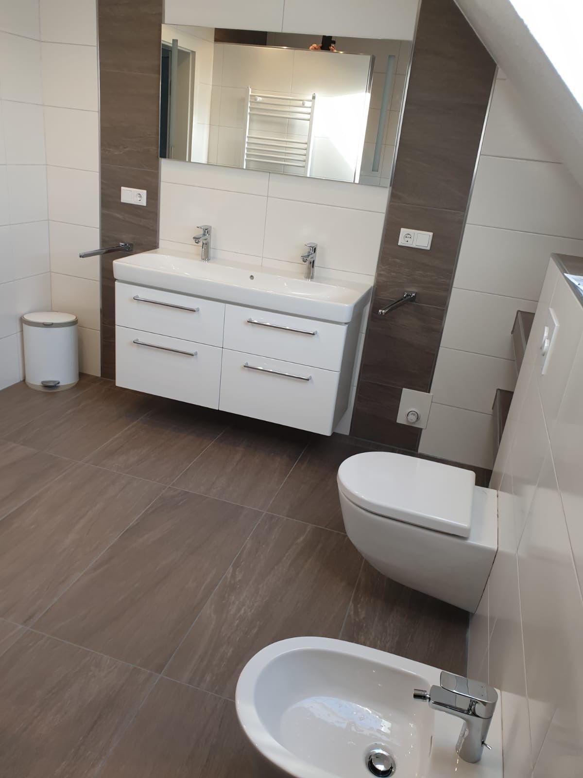 Modernes Badezimmer mit freischwebendem Doppelwaschbecken, WC, Bidet und braunem Fliesenboden. Weiße und braune Wände.