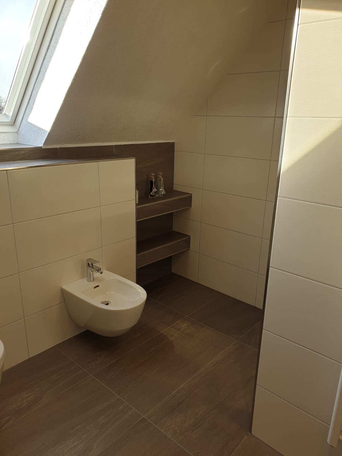 Badezimmer mit weißen Fliesen, Bidet und eingebautem Regal. Brauner Boden und braune Wandakzente.