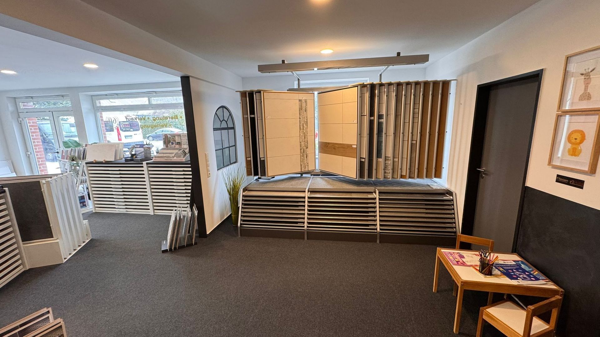 Innenansicht eines Geschäfts, in dem Bodenbelagsmuster ausgestellt sind. Regale, Schaufenster, Tür und ein Tisch mit Stuhl