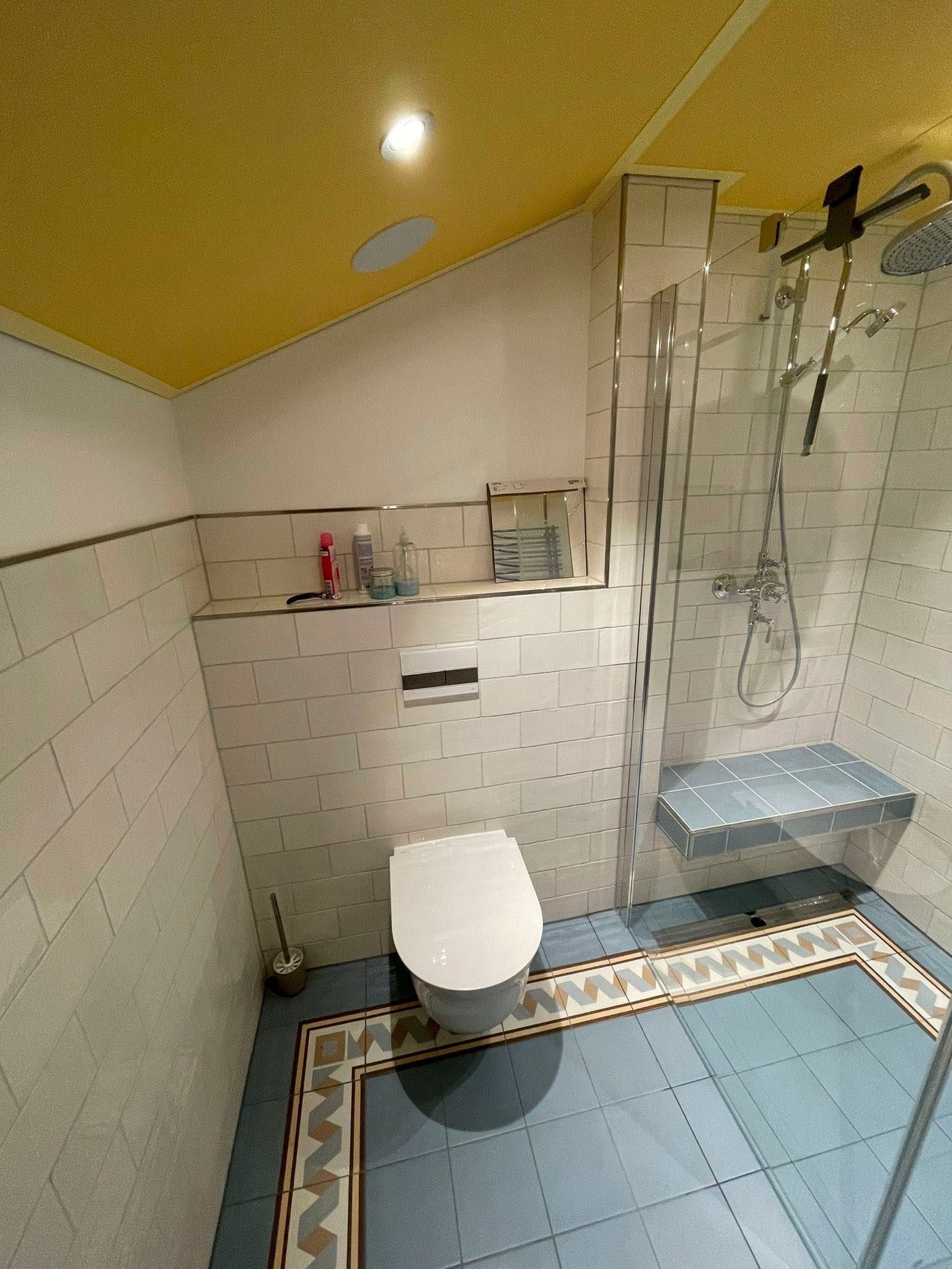 Badezimmer mit blau gefliestem Boden, weißer Toilette und Glasdusche. Gelbe Decke.
