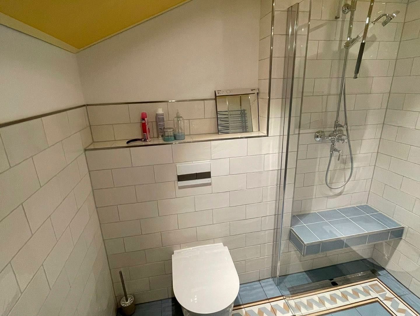 Badezimmer mit weiß gefliesten Wänden, Toilette, Dusche mit Glastür und einer blauen Sitzbank. Gelbe Decke.