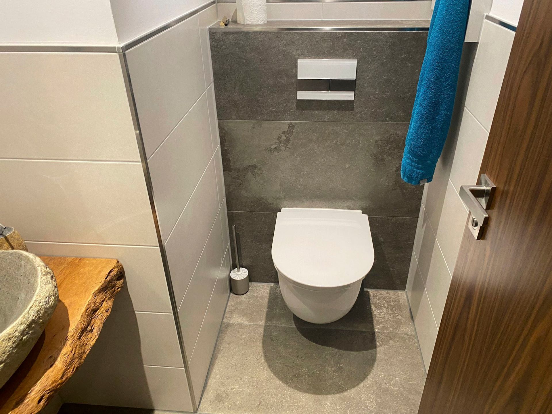 Toilette in einem kleinen Badezimmer. Graue und weiße Fliesen, Holztür und ein blaues Handtuch.