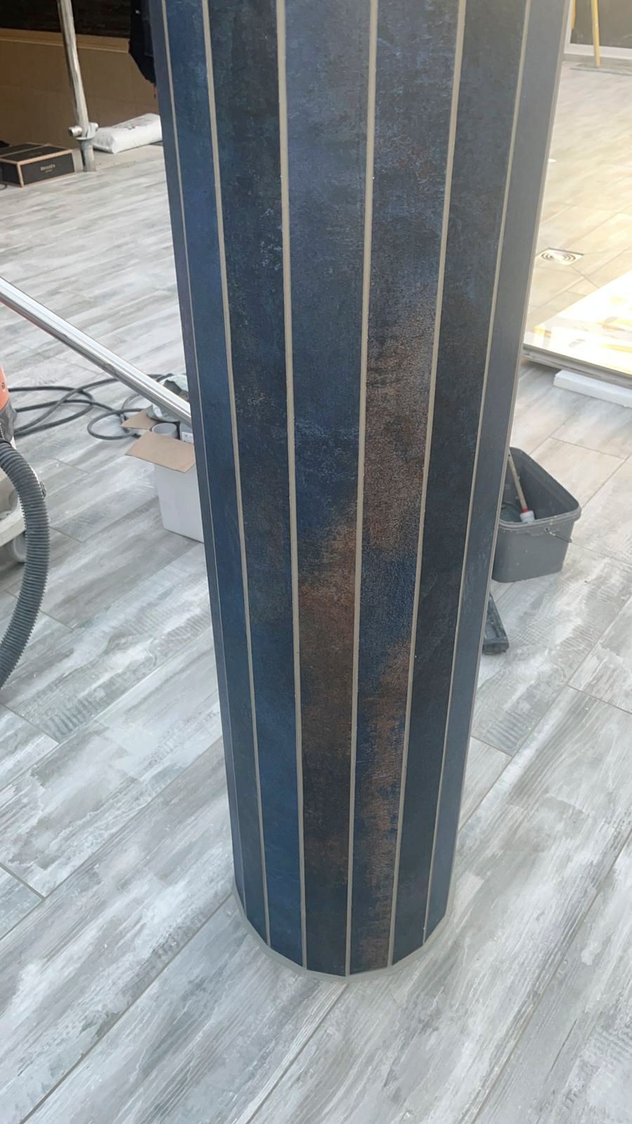 Eine große, zylindrische Rolle aus schwarzem Material mit vertikalen hellbraunen Streifen steht auf einer gefliesten Fläche.