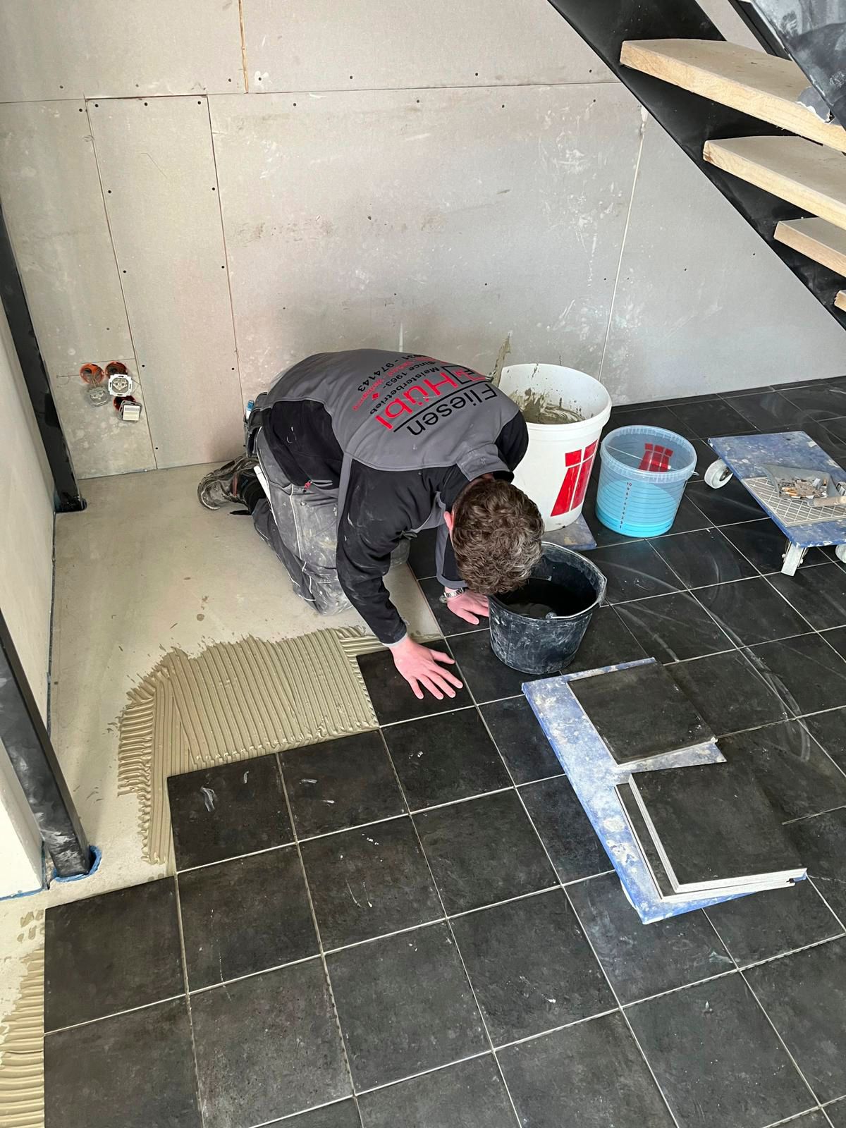 Eine Person verlegt Fliesen auf einem Boden und bringt dunkle Fliesen mit einer Kelle an