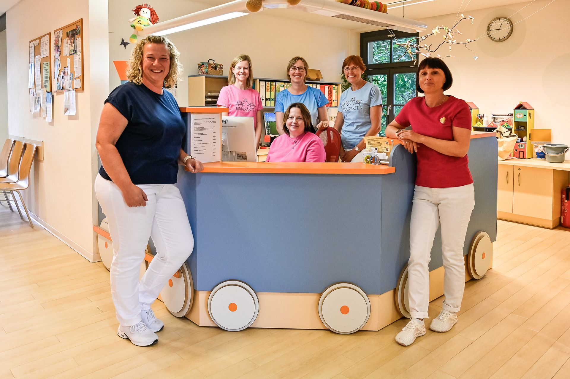 Eine Gruppe von sieben Personen steht hinter einem blau-orangenen Empfangstresen in einem Büro.