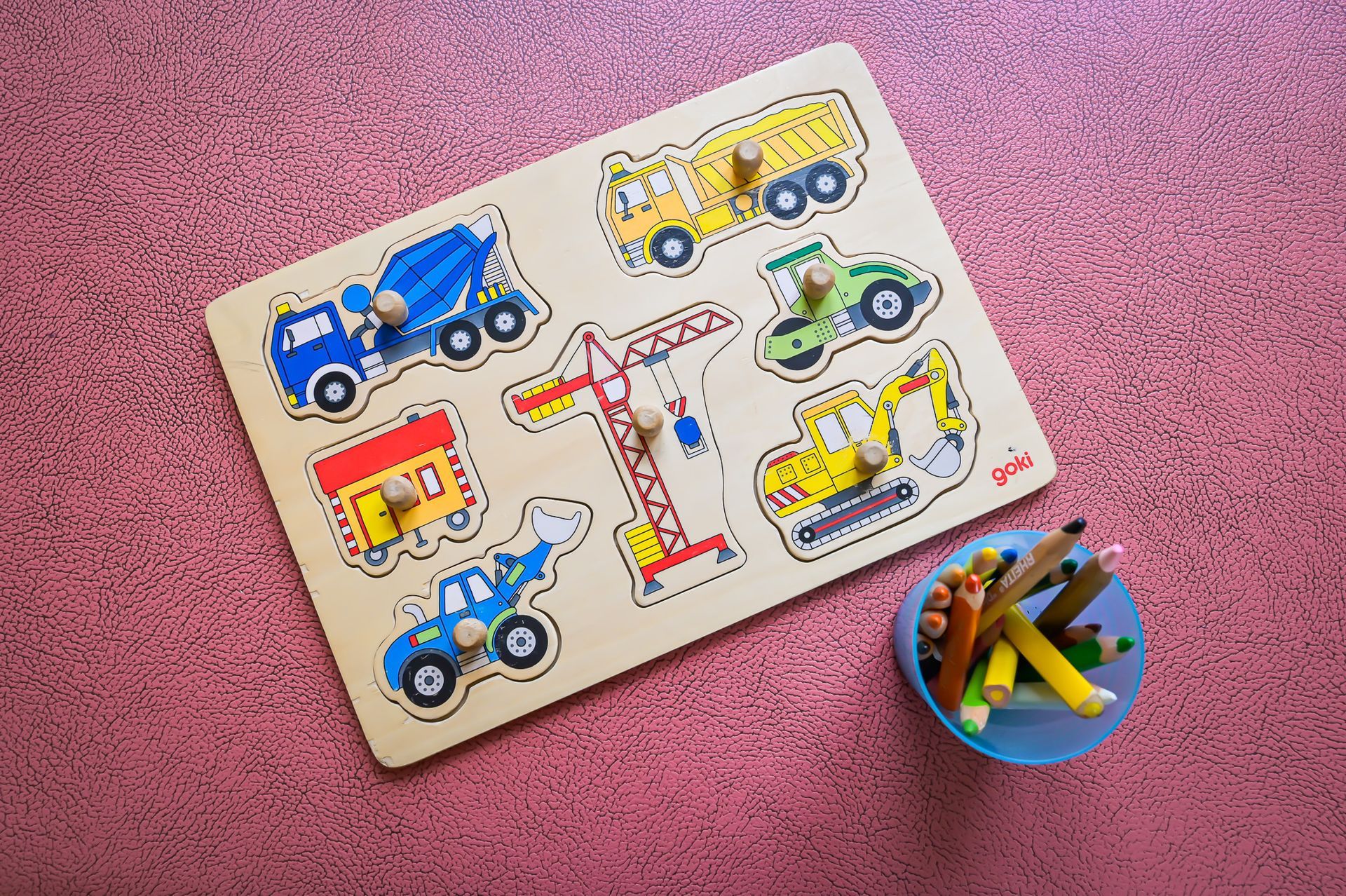 Holzpuzzle mit Baustellenfahrzeugen; daneben stehen Stifte in einem blauen Becher auf einem rosa Teppich.