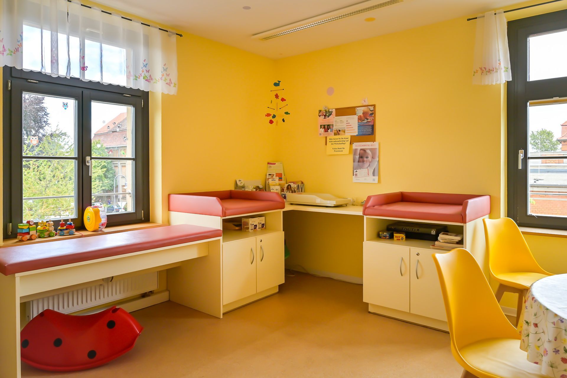 Ein gelbes Zimmer mit Wickeltischen, einem Fenster und gelben Stühlen.