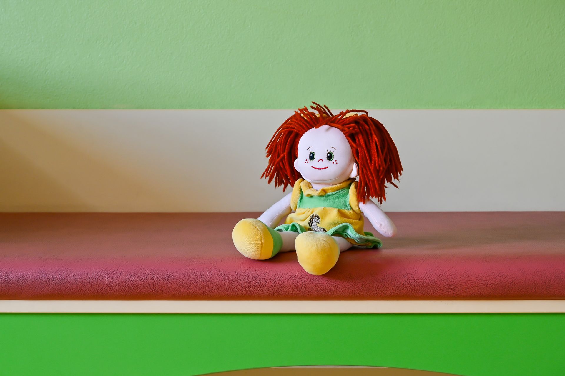 Puppe mit roten Haaren und gelbem Kleid sitzt auf einem rosa Sims vor einer grünen Wand.