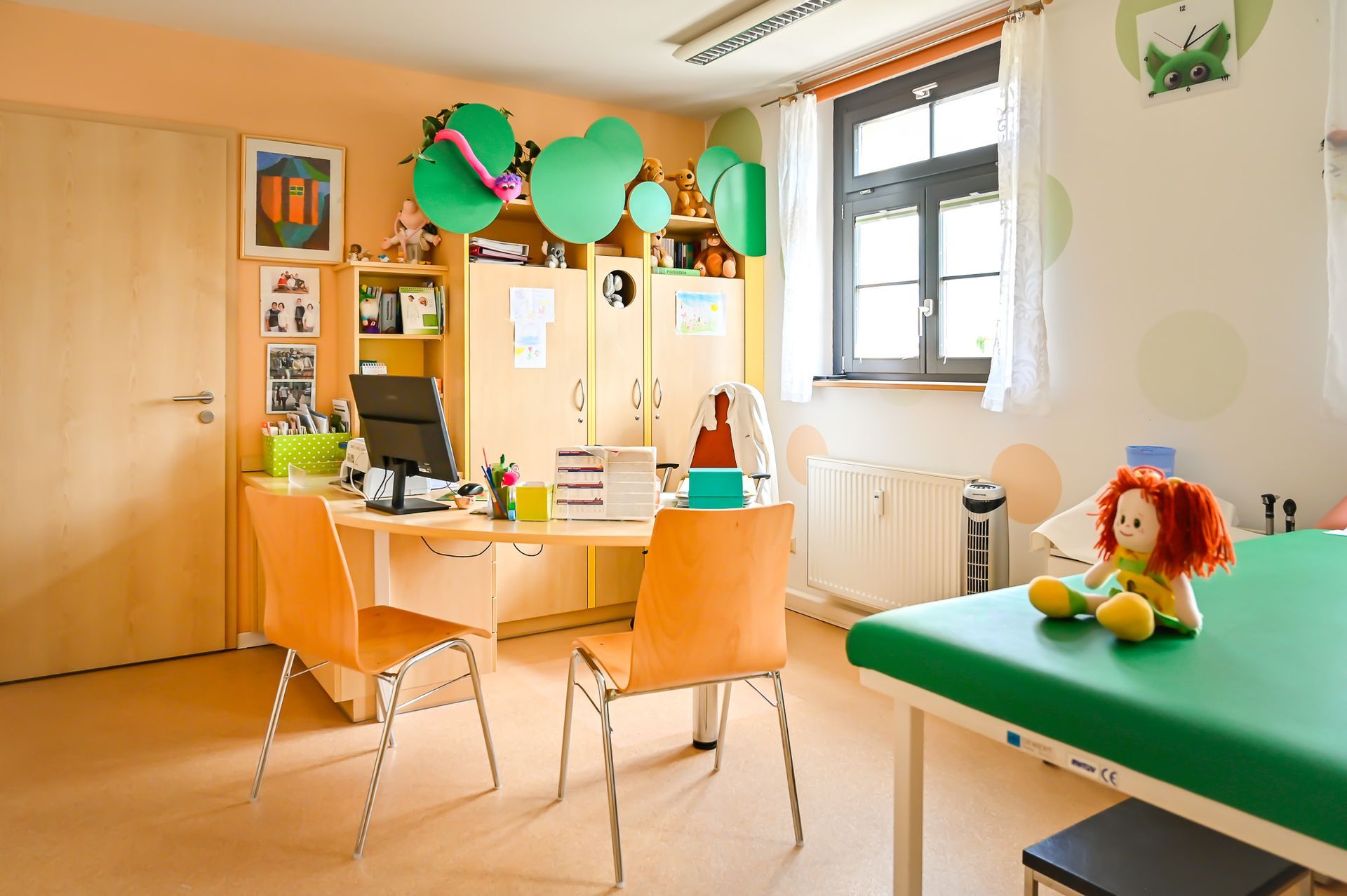 Arztpraxis: Schreibtisch, Stühle, Untersuchungsliege, Spielzeug. Hellorange Wände, grüne Akzente.