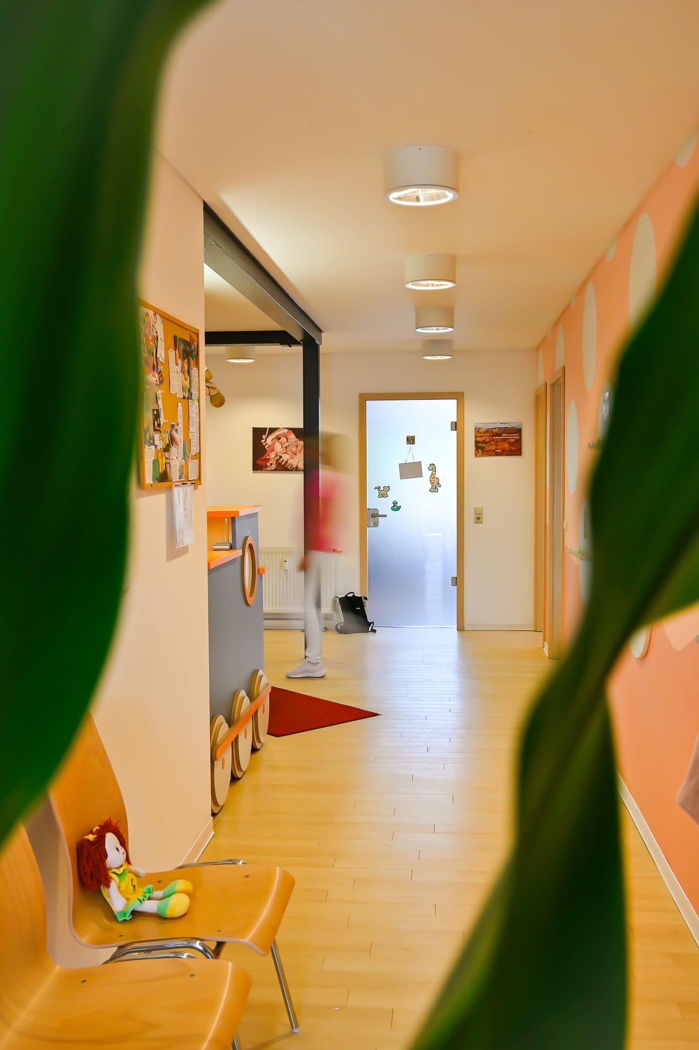 Langer Flur mit Holzboden, Pflanze im Vordergrund, Person im Hintergrund verschwommen, und Türen.