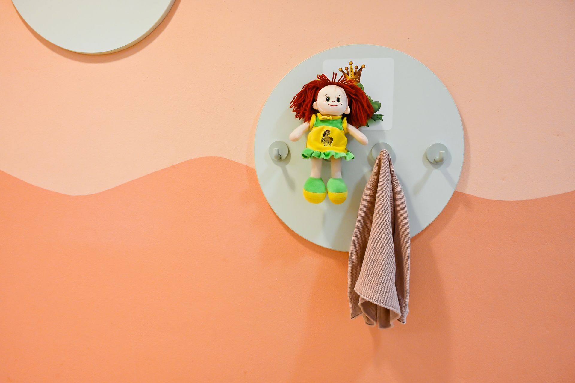 Puppe und Handtuch hängen an einer runden, hellgrauen Wandhalterung vor einer pfirsichfarben und weiß gestrichenen Wand.