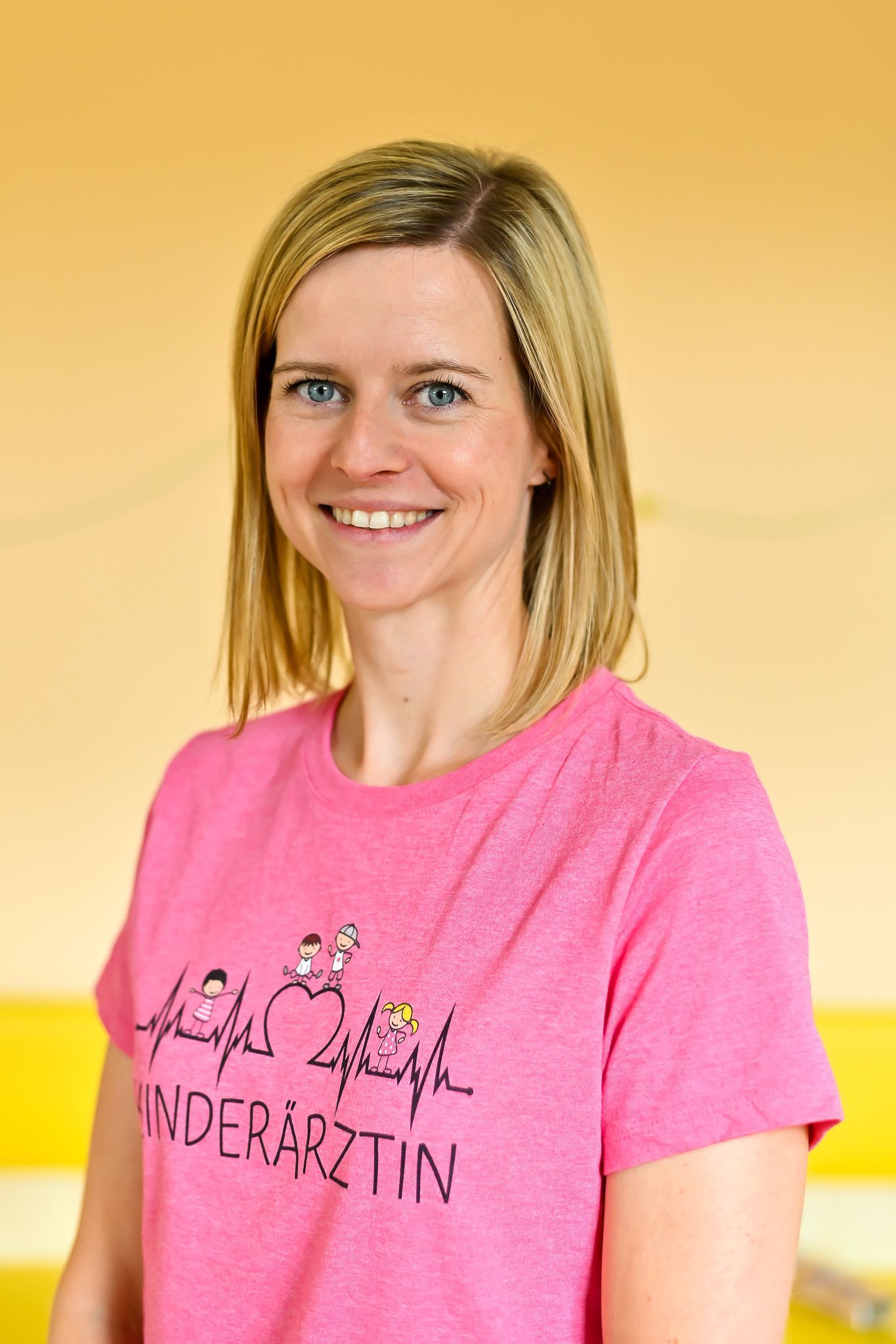 Eine Frau in einem rosa Hemd mit der Aufschrift „Kinderärztin“ lächelt.