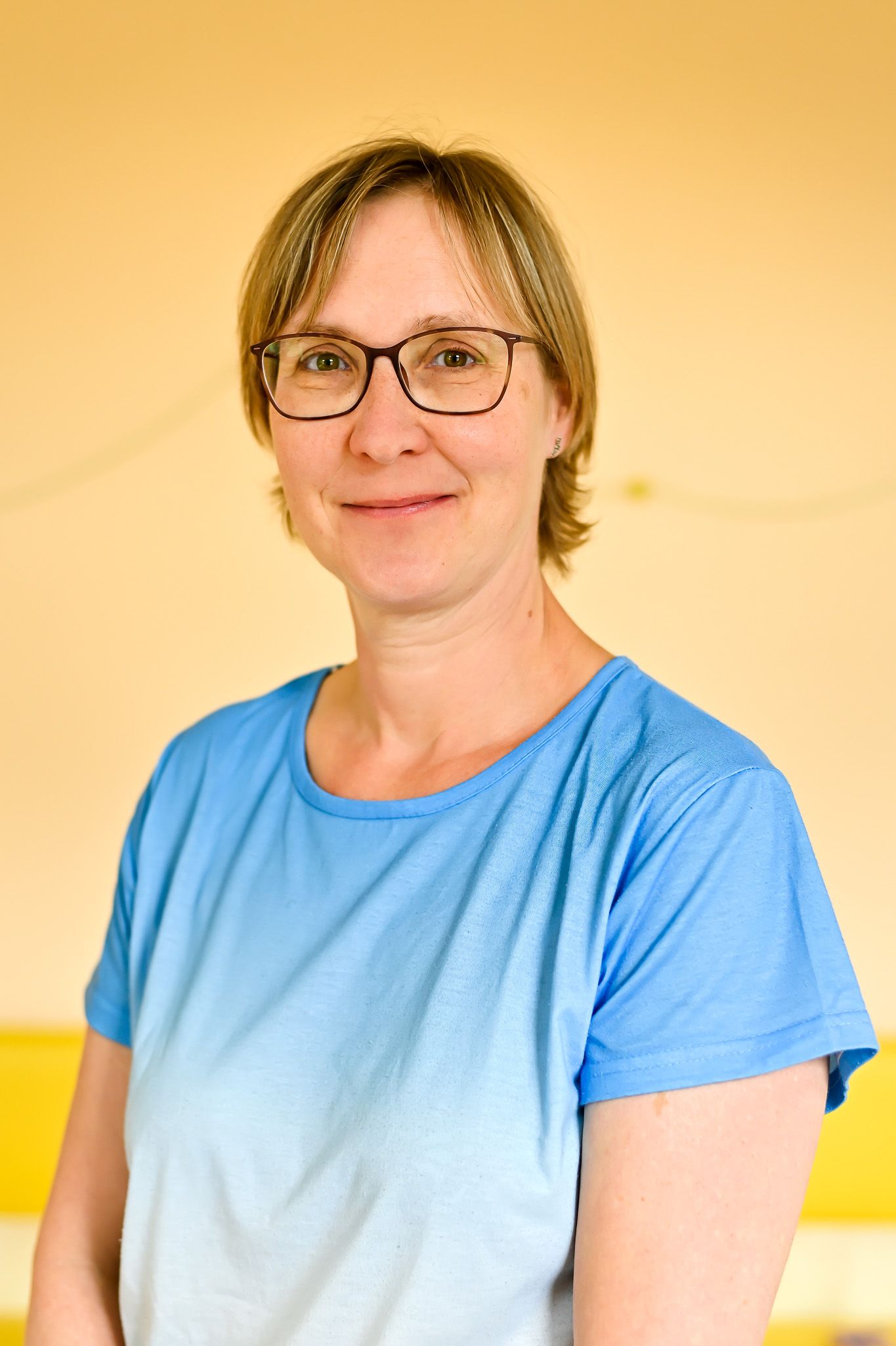 Eine Frau mit Brille und blauem Hemd lächelt vor einer gelben Wand.