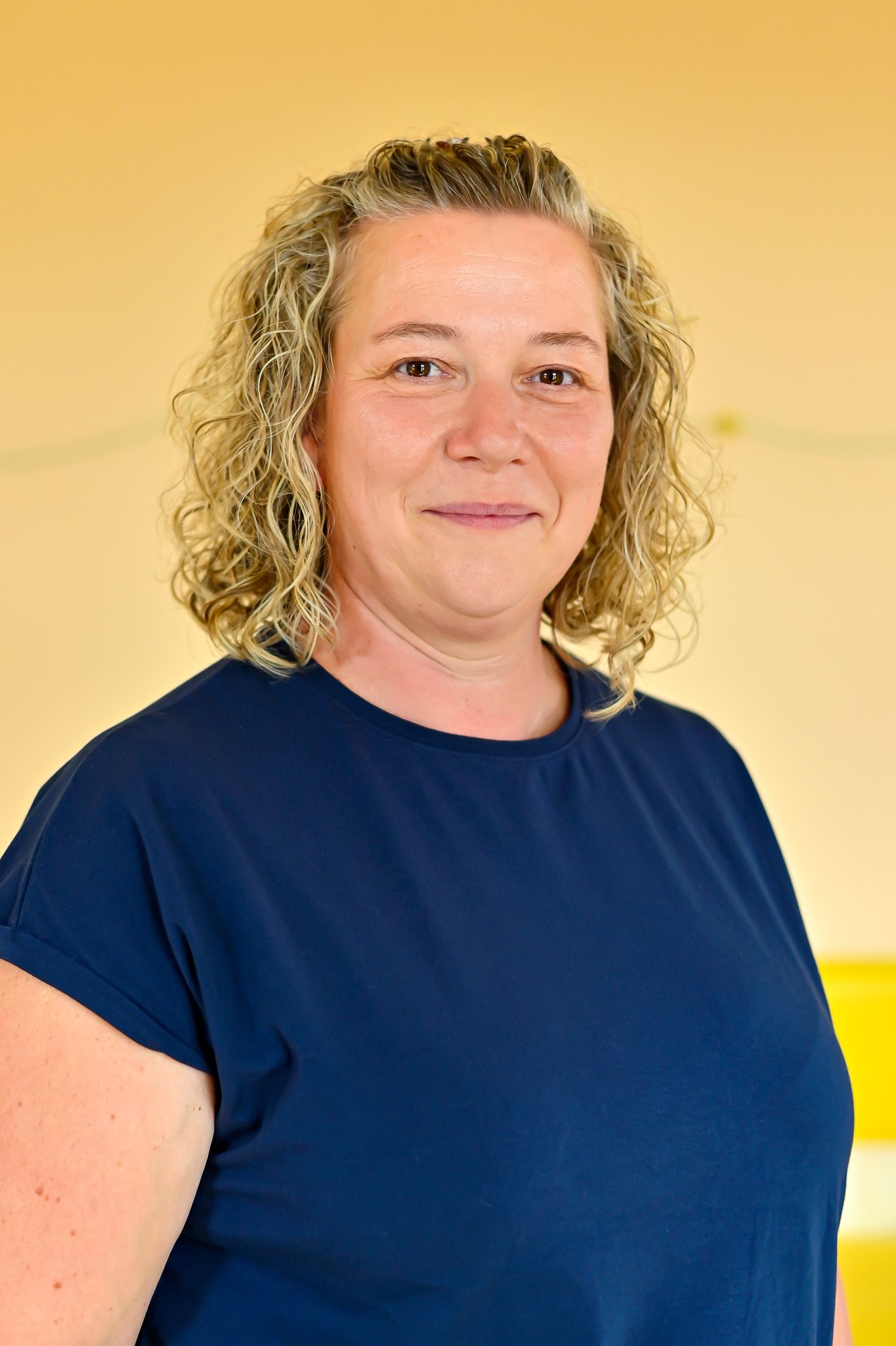 Eine Frau mit lockigem Haar und einem blauen Oberteil lächelt vor einer gelben Wand.