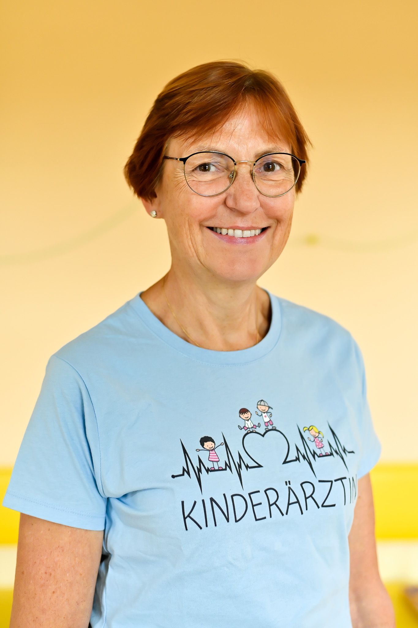 Frau mit Brille, die ein hellblaues T-Shirt mit der Aufschrift „Kinderarzt“ trägt, lächelt vor einem gelben Hintergrund.